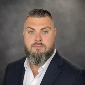 Professional headshot of John A. Hamilton wearing a dark suit jacket and white shirt against a neutral gray background.